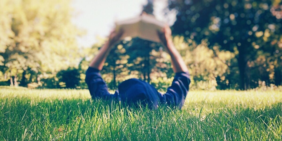 Persona tirada en el césped artificial leyendo un libro con sus brazos en lo alto